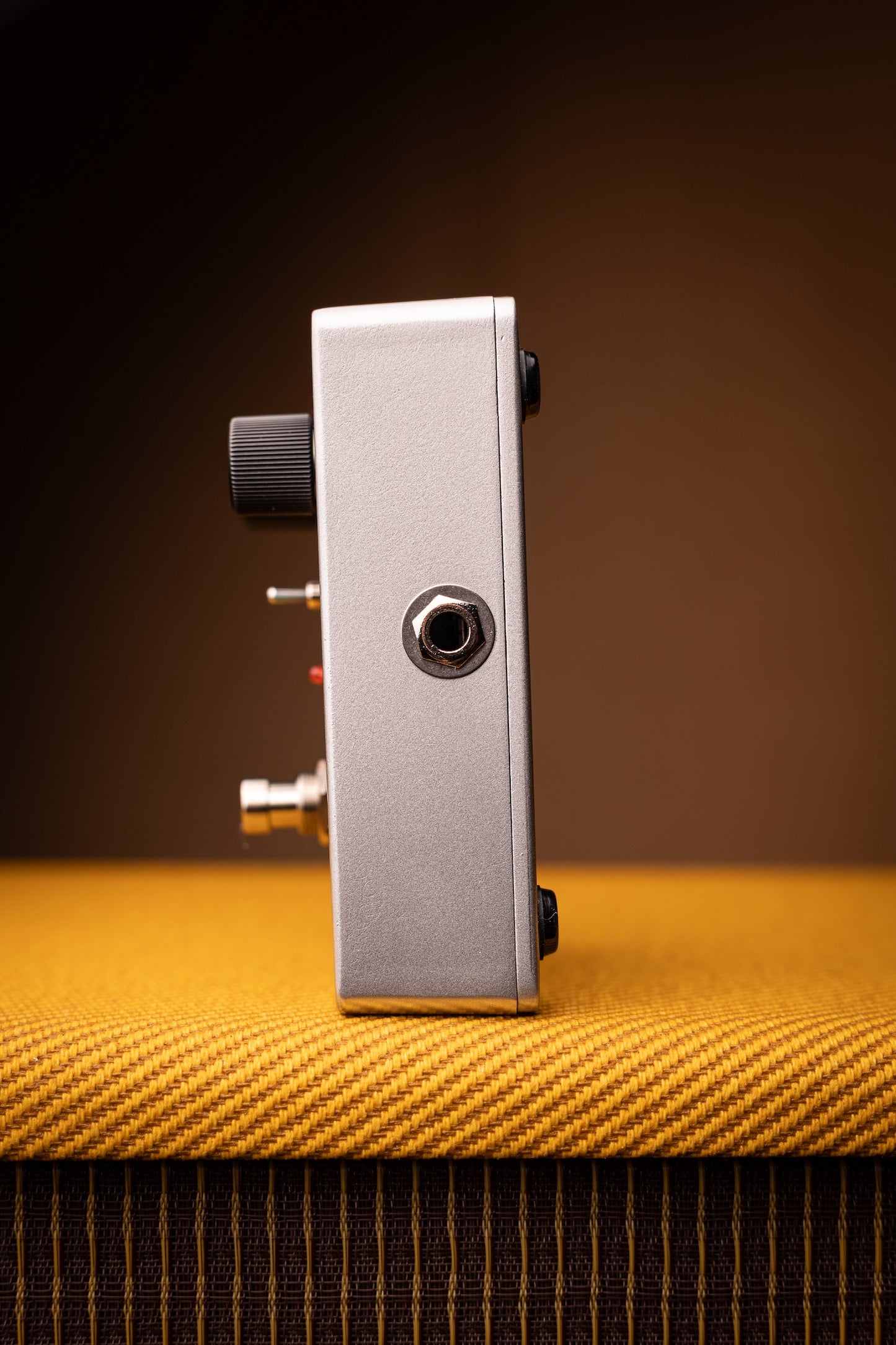A silver metal guitar effects pedal with black knobs stands upright on a textured yellow-orange surface against a dark brown background.