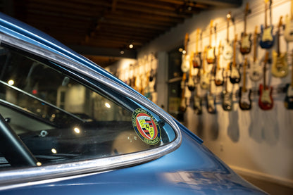Close-up of a car's window with a Porsche logo, blurred background of guitars on a wall.
