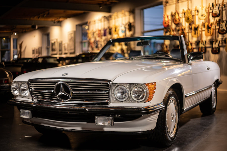 A 1989 Mercedes-Benz 560 SL car in Arctic White color, with visible blue leather interior, parked inside a showroom with various guitars hanging on the walls.