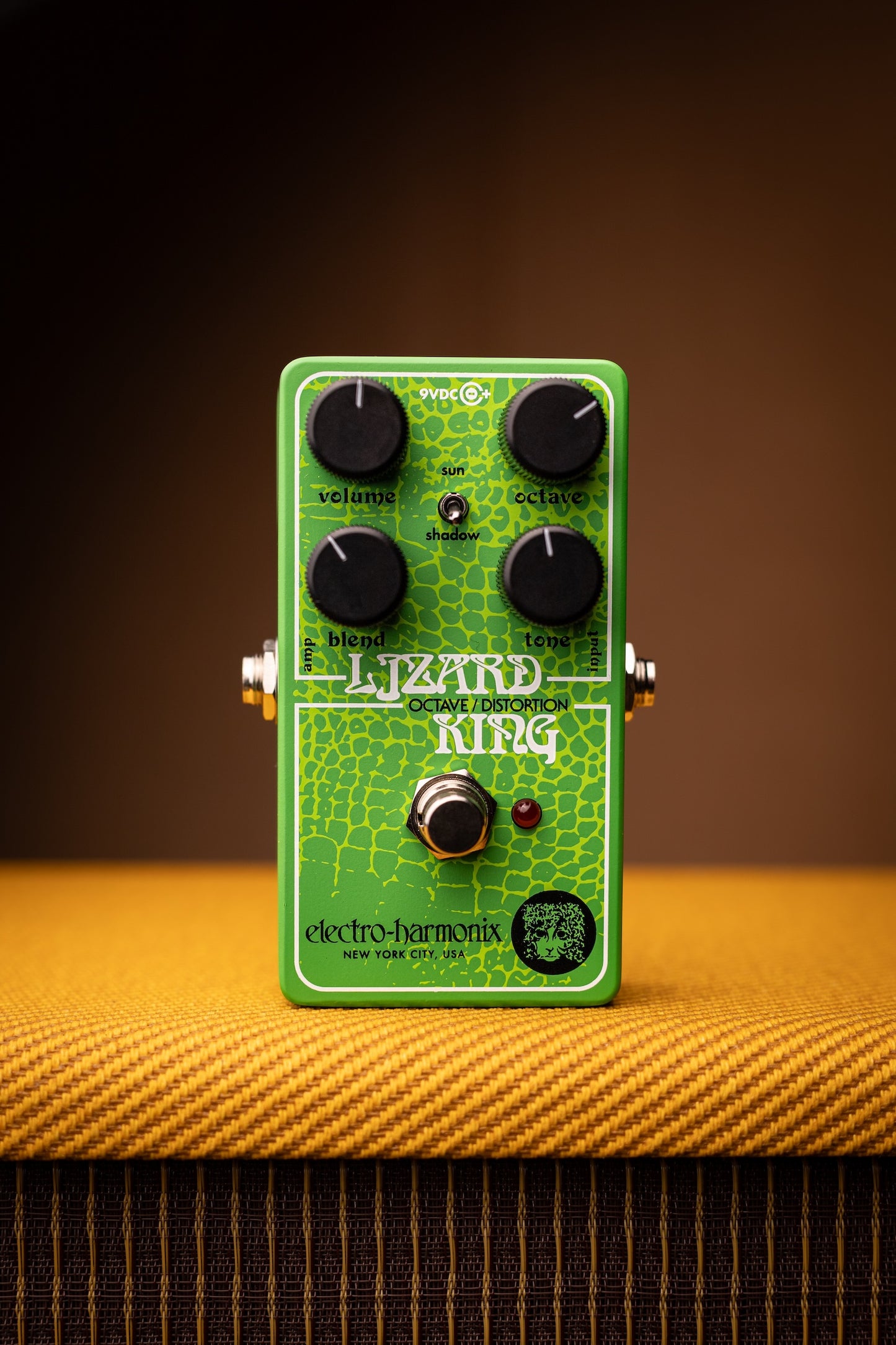 A green Electro-Harmonix Lizard King overdrive guitar pedal with black knobs, standing on a textured yellow-orange surface against a dark brown background.