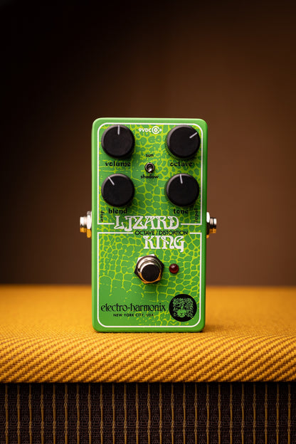 A green Electro-Harmonix Lizard King overdrive guitar pedal with black knobs, standing on a textured yellow-orange surface against a dark brown background.