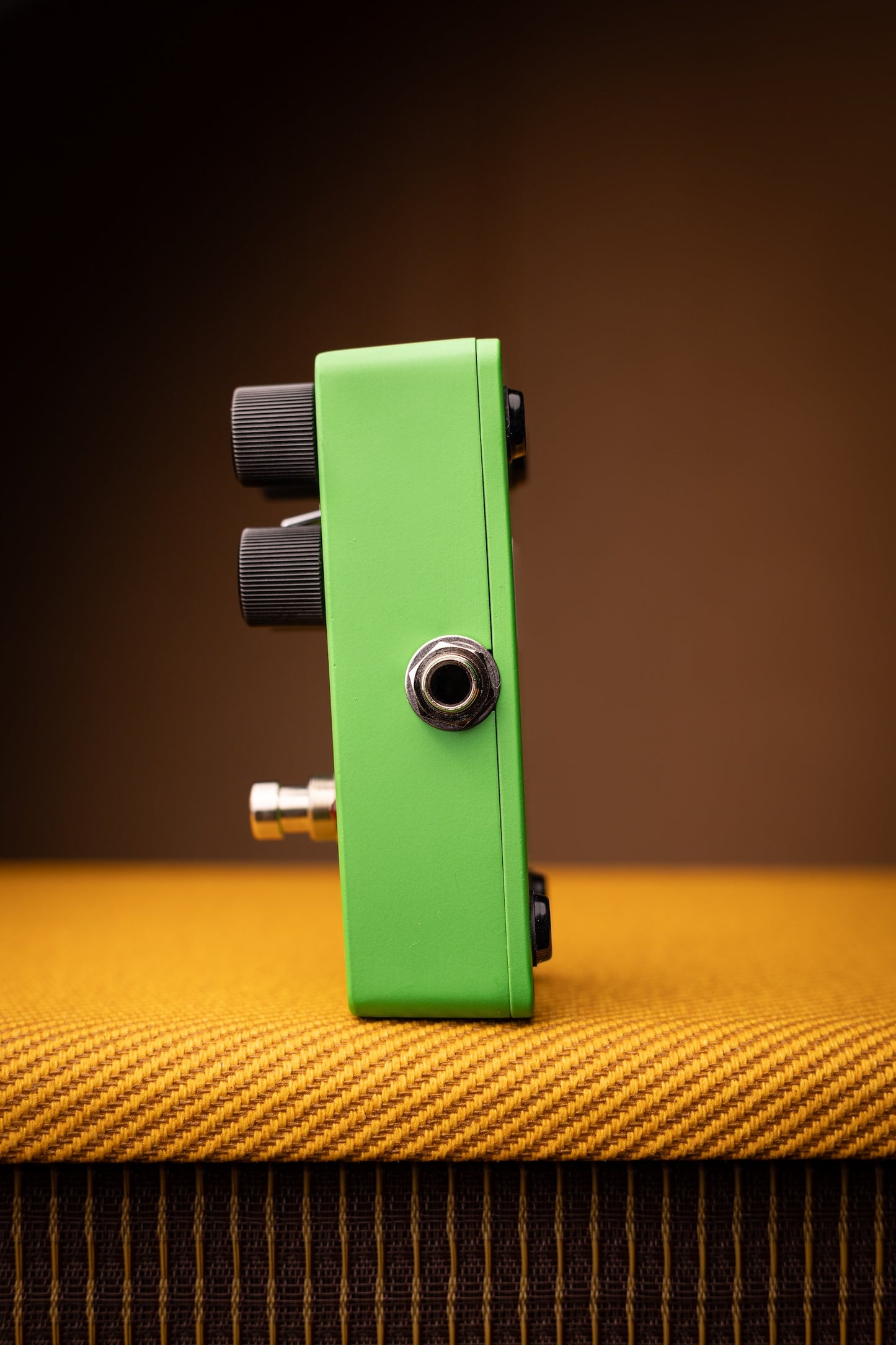 A bright green guitar effects pedal with black knobs and a silver metal jack input, standing upright on a textured yellow-orange surface against a dark brown background.