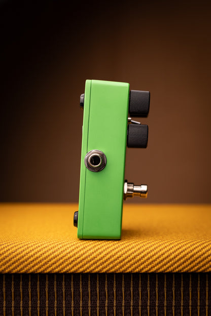 A green guitar effects pedal with black knobs and a silver jack connector, standing upright on a textured yellow-orange surface against a dark brown background.