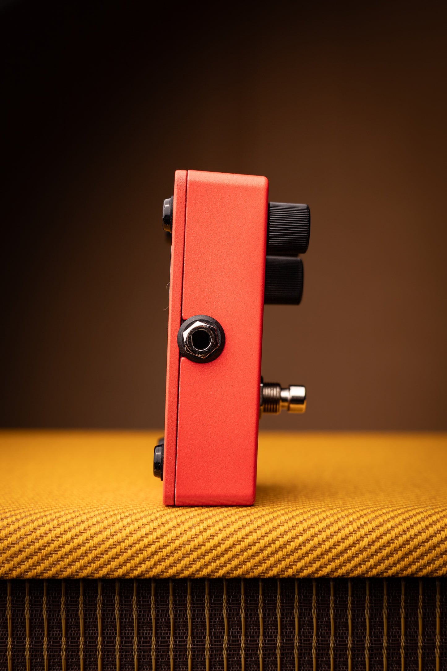 A red guitar effects pedal with black knobs and a gold-toned jack connector, standing upright on a textured yellow surface against a dark brown background.