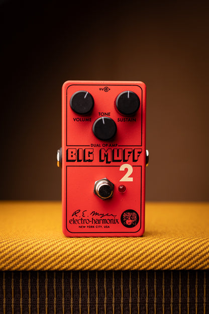 An Electro-Harmonix Big Muffl 2 guitar effects pedal in red, resting on a yellow guitar headstock fabric cover against a dark brown background.