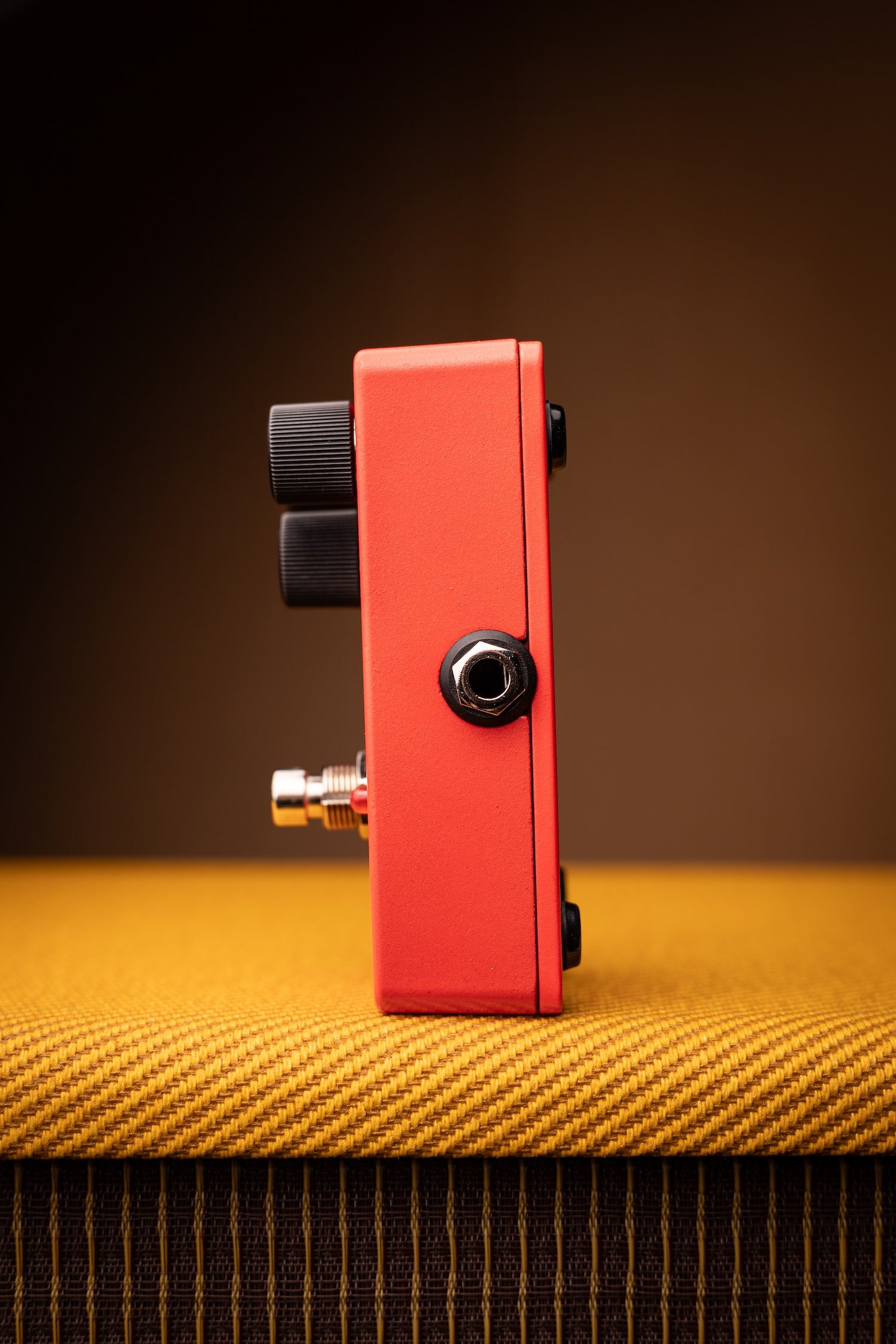 A red guitar effects pedal with black knobs and a silver jack input, standing upright on a yellow textured surface against a dark brown background.