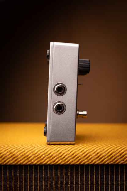 A silver metal guitar effects pedal with black knobs and jacks, standing upright on a textured yellow-orange surface against a dark brown background.