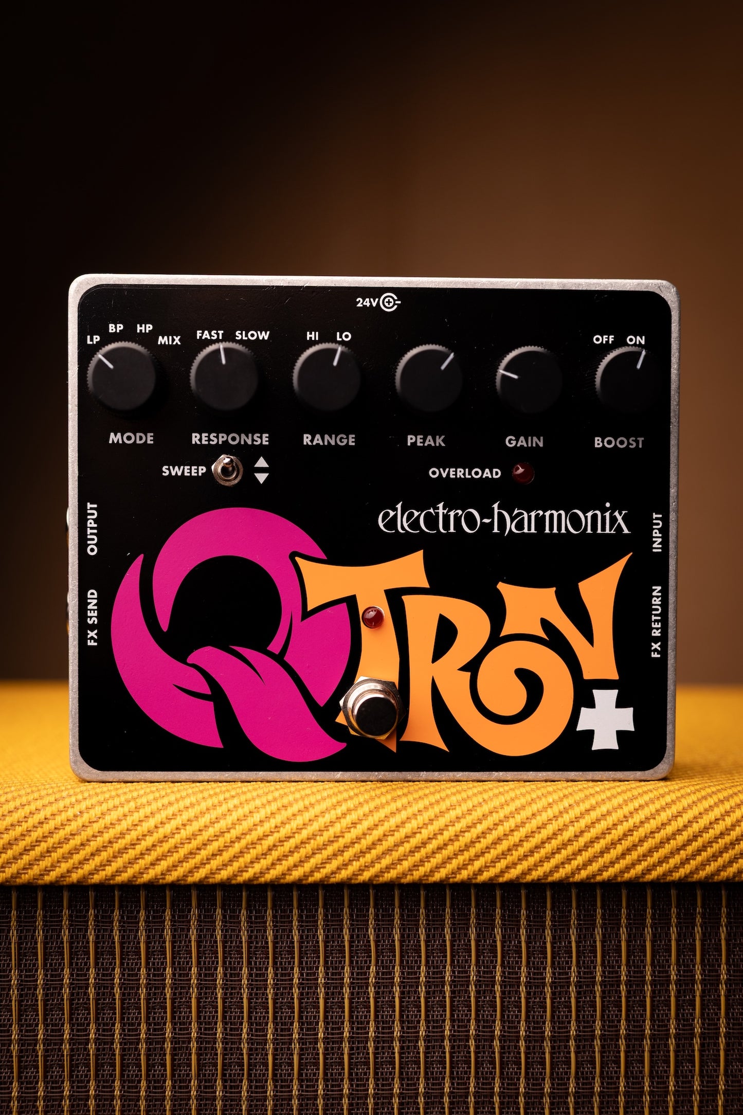 Electro-Harmonix QTRN+ guitar effects pedal with black faceplate, silver metal edges, and pink and orange graphic lettering, resting on a yellow guitar headstock.