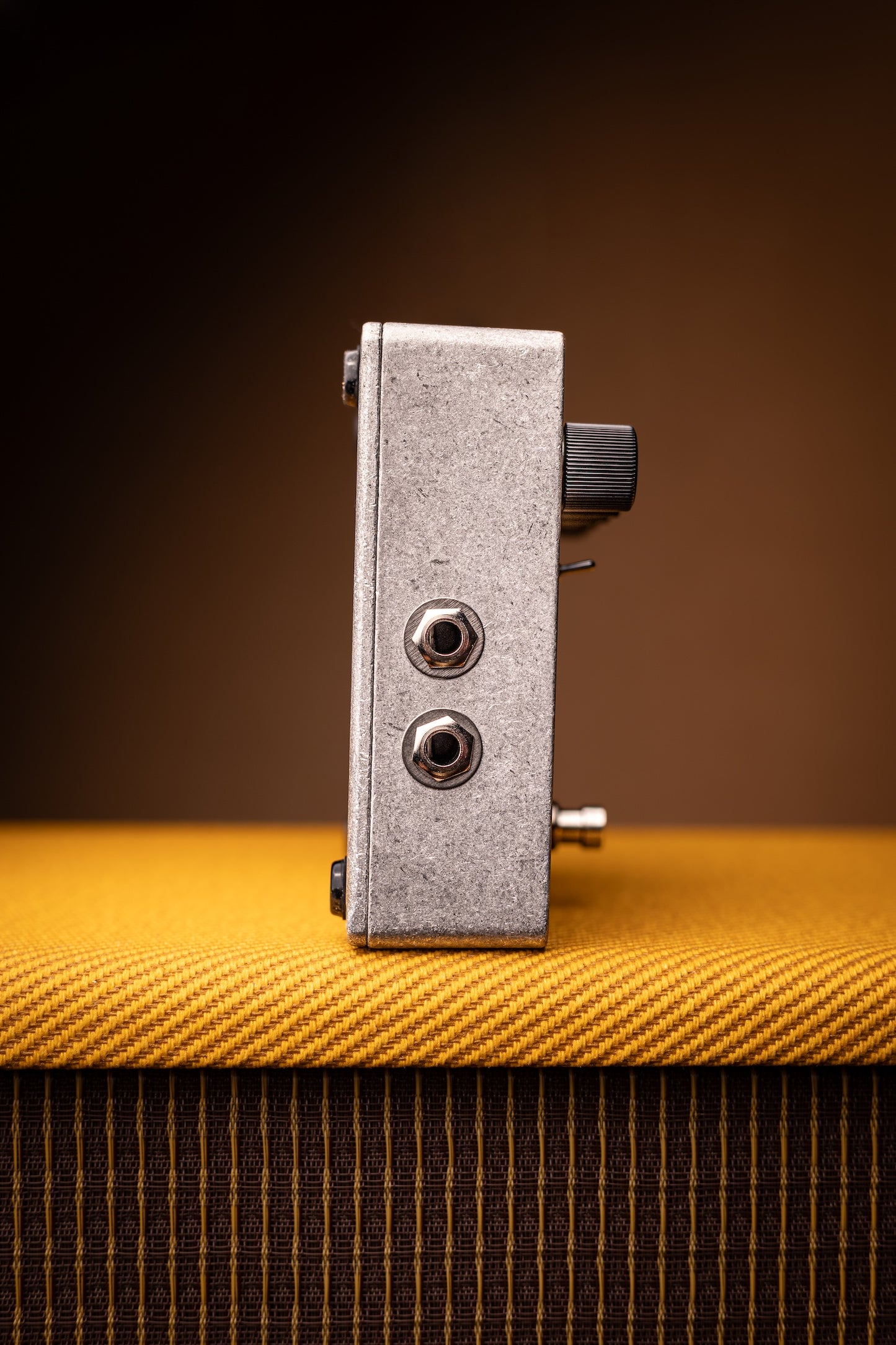 A silver metal guitar effects pedal with black knobs, standing upright on a textured yellow fabric surface against a dark brown background.