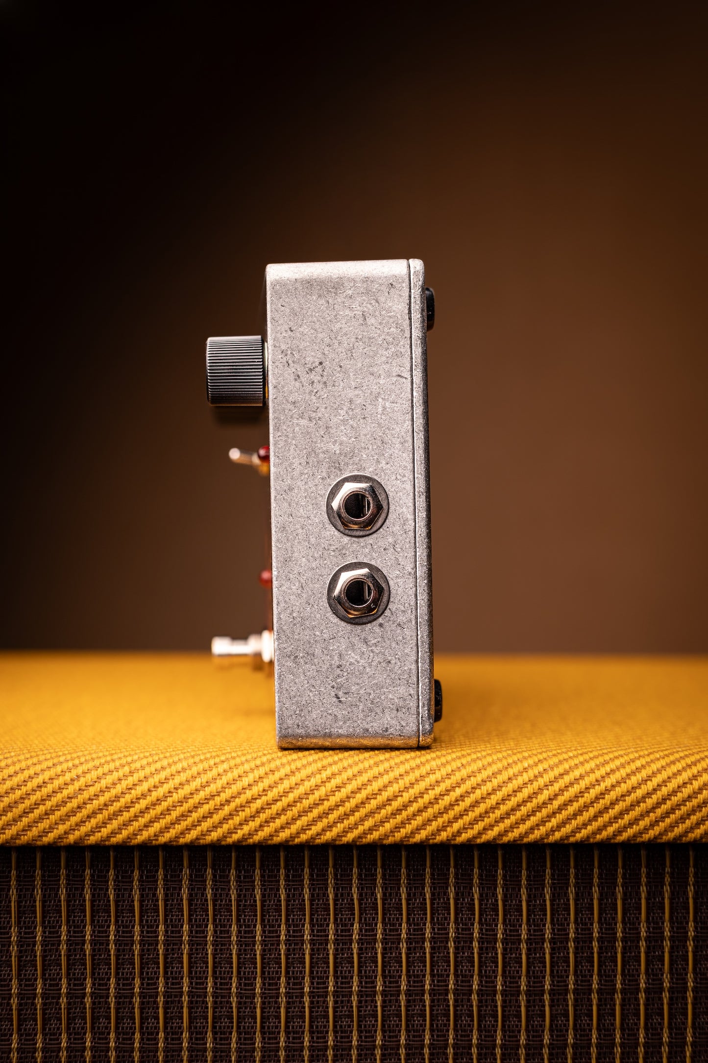 A small rectangular metal guitar effects pedal with two jacks and a black knob, standing upright on a yellow textured surface against a dark brown gradient background.