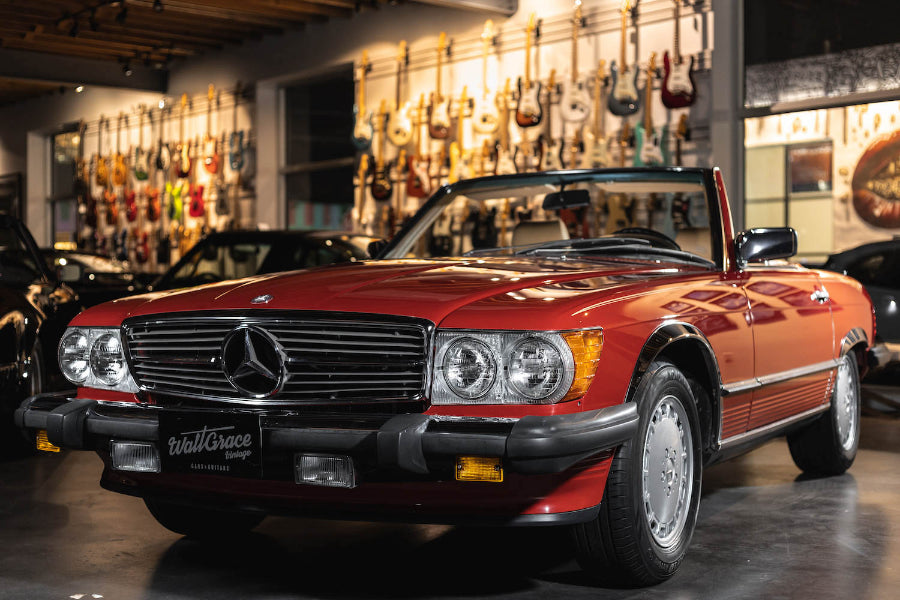 A red 1989 Mercedes-Benz 560 SL car parked inside a showroom with various other cars and musical instruments in the background.