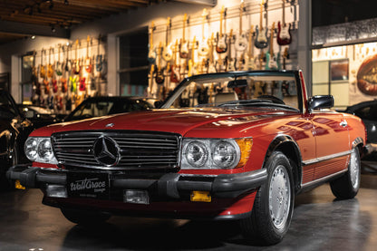 A red 1989 Mercedes-Benz 560 SL car parked inside a showroom with various other cars and musical instruments in the background.