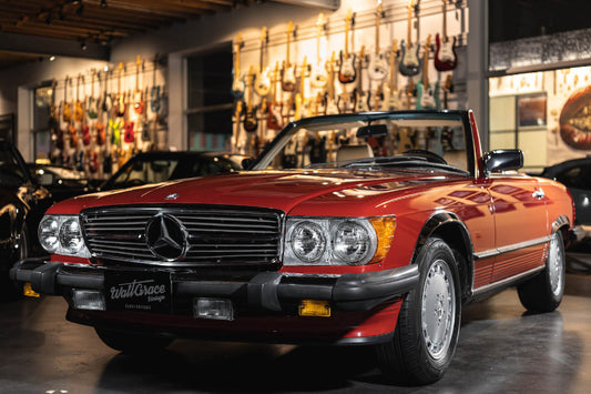 A red 1989 Mercedes-Benz 560 SL car parked inside a showroom with various other cars and musical instruments in the background.