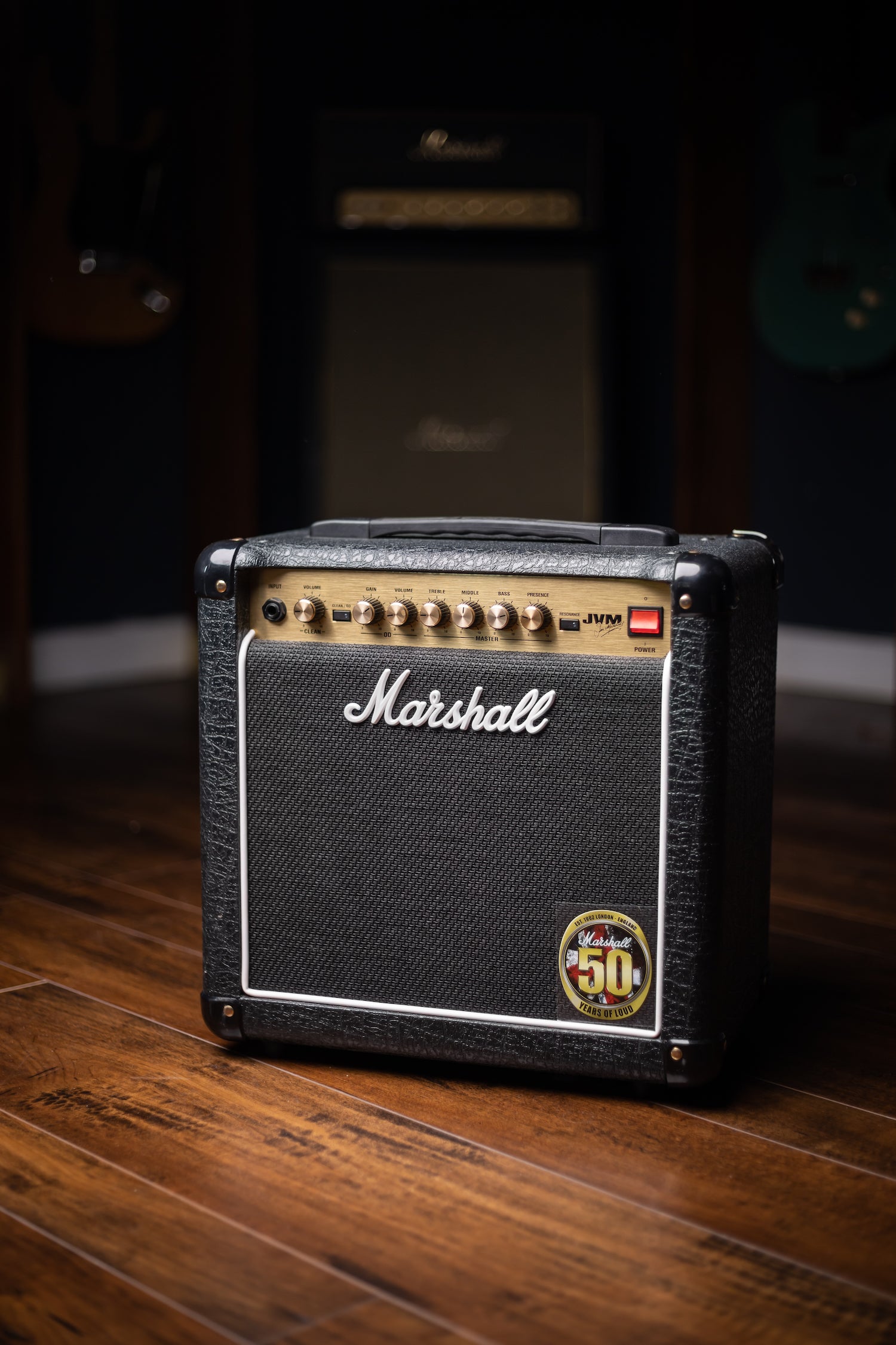 A black Marshall JVM Combo Amp with silver detailing on a wooden floor in a room with musical instruments in the background.